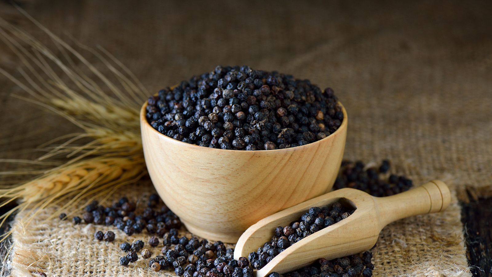 Wooden bowl and scoop filled with black peppers on burlap.