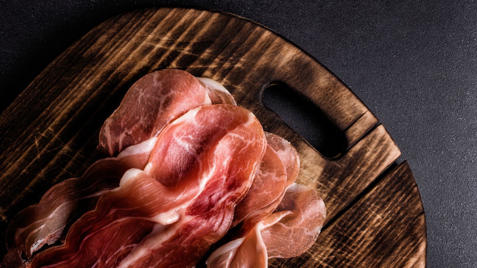 Thin slices of cured ham on a wooden board with dark backdrop.