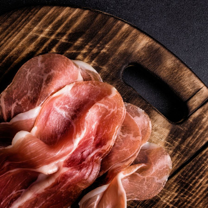 Thin slices of cured ham on a wooden board with dark backdrop.