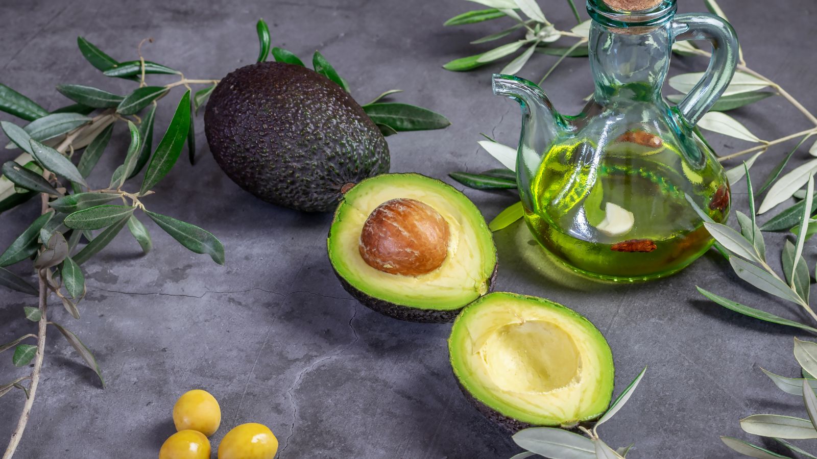 Whole and sliced avocado with avocado oil in a glass jar on gray surface.