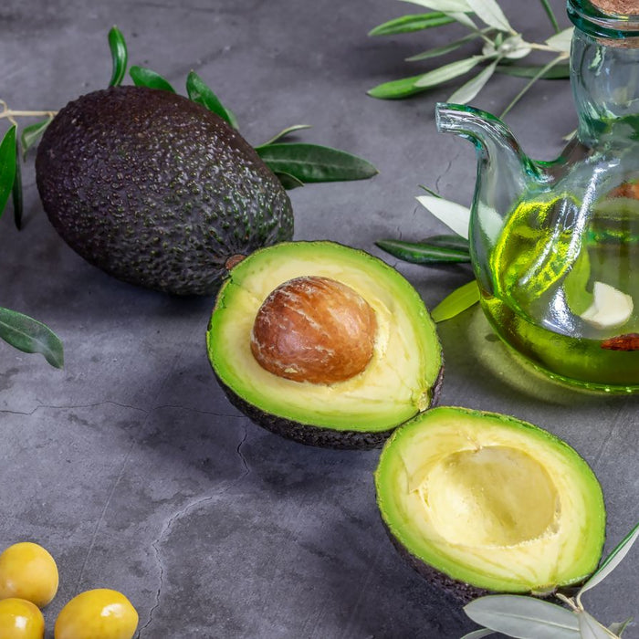 Whole and sliced avocado with avocado oil in a glass jar on gray surface.