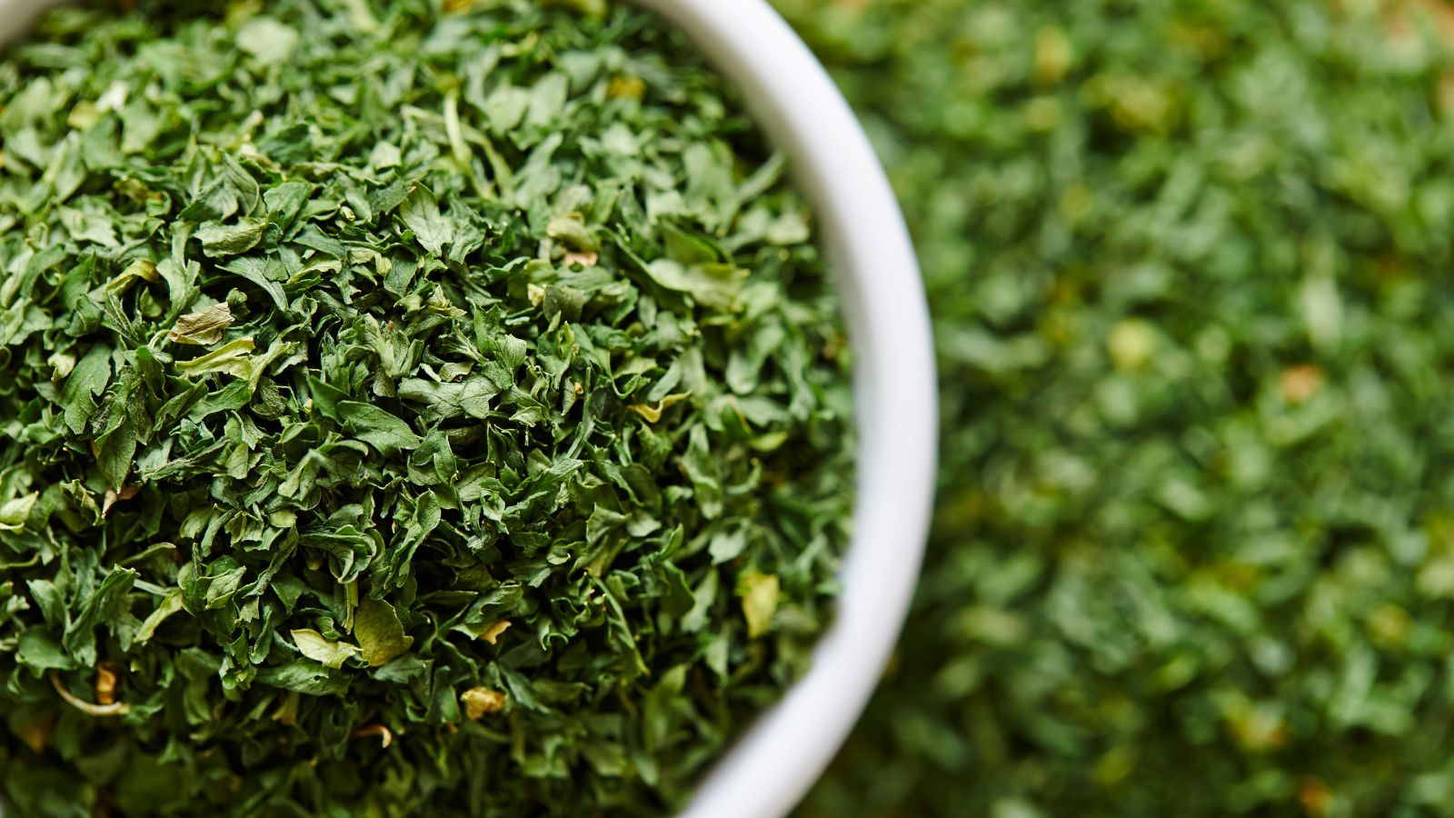 Close-up of dried parsley flakes in white bowl on green background.