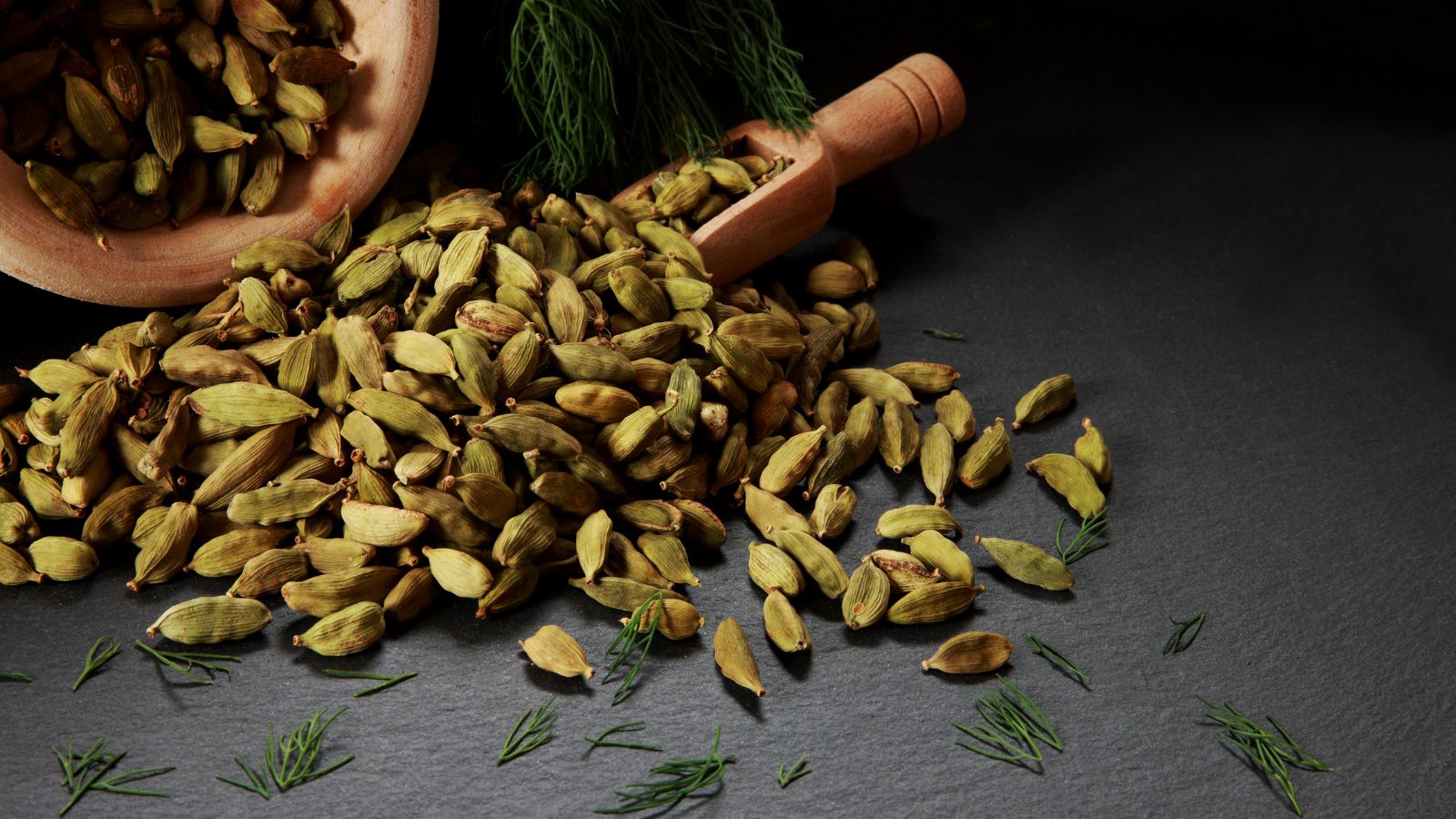 Cardamom pods spilled from a wooden bowl with herbs on a dark surface.