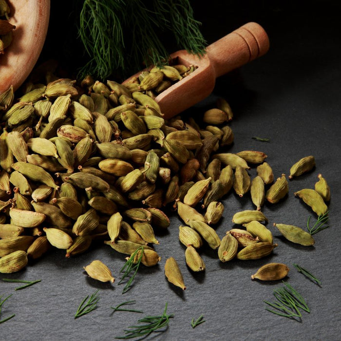 Cardamom pods spilled from a wooden bowl with herbs on a dark surface.