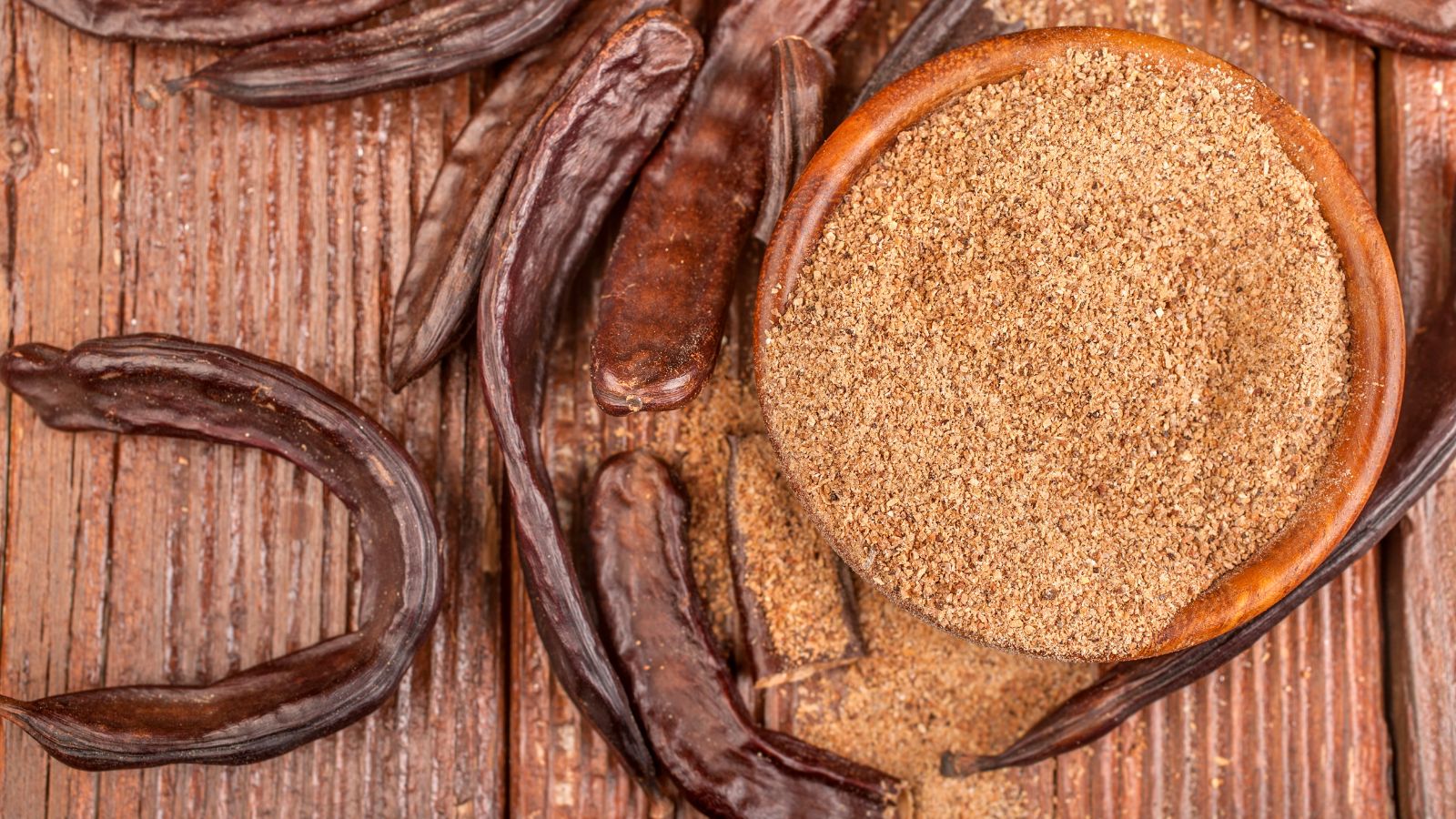 Ground carob powder in a wooden bowl with dried carob pods on a table.