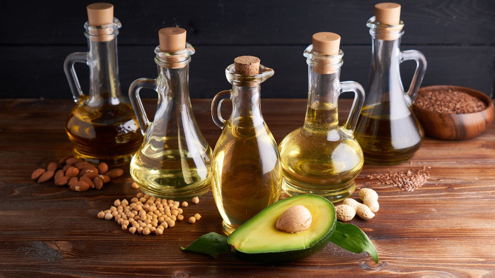 Assorted vegetable oils in glass bottles with avocado on wooden table.