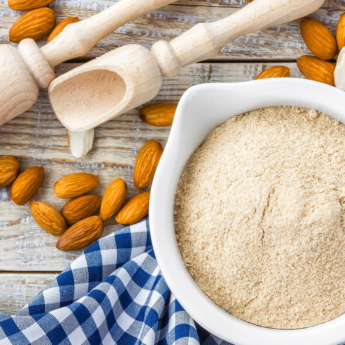 Wooden scoops and raw almonds surround a bowl filled with almond flour.