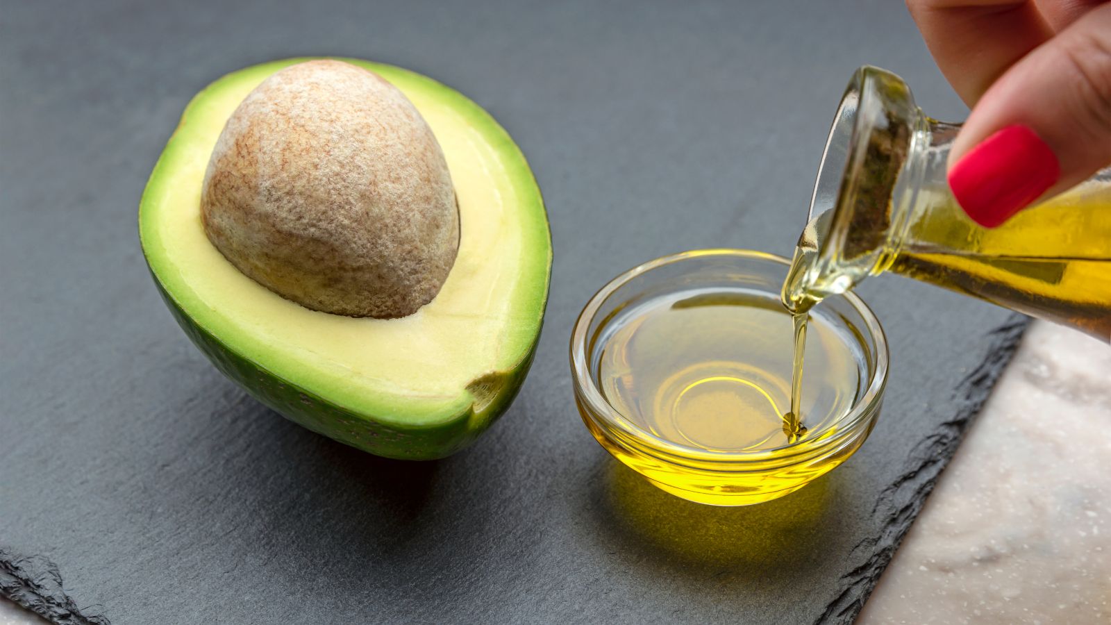Avocado half with seed and avocado oil being poured in a glass bowl.