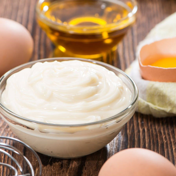 Fresh creamy mayo in glass bowl with eggs and oil on table.