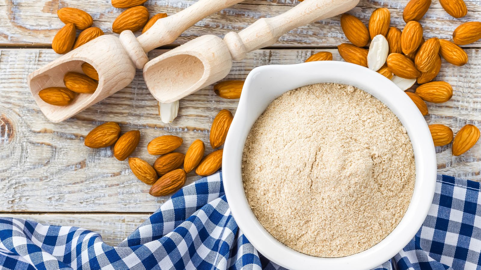 Wooden scoops and raw almonds surround a bowl filled with almond flour.