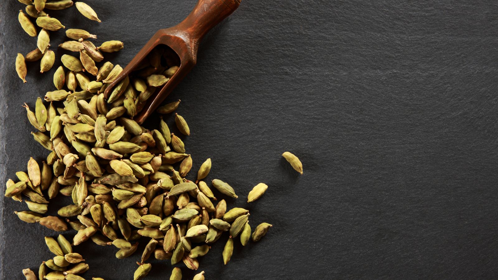 Cardamom pods scattered on a dark slate background with a scoop.