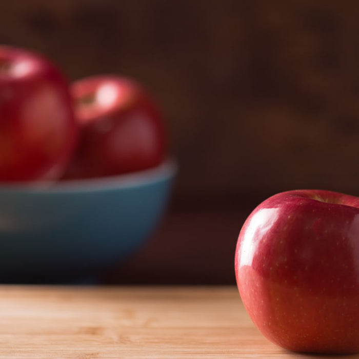 Shiny red apple on wooden surface with bowl of apples in background.