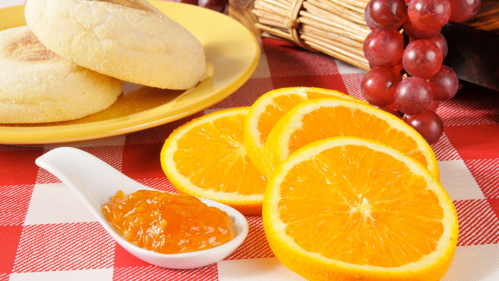 Orange slices with English muffins and marmalade on a red tablecloth.