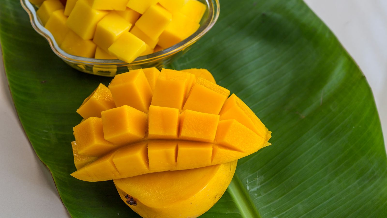 Diced mango cubes in glass bowl on large green banana leaf surface.