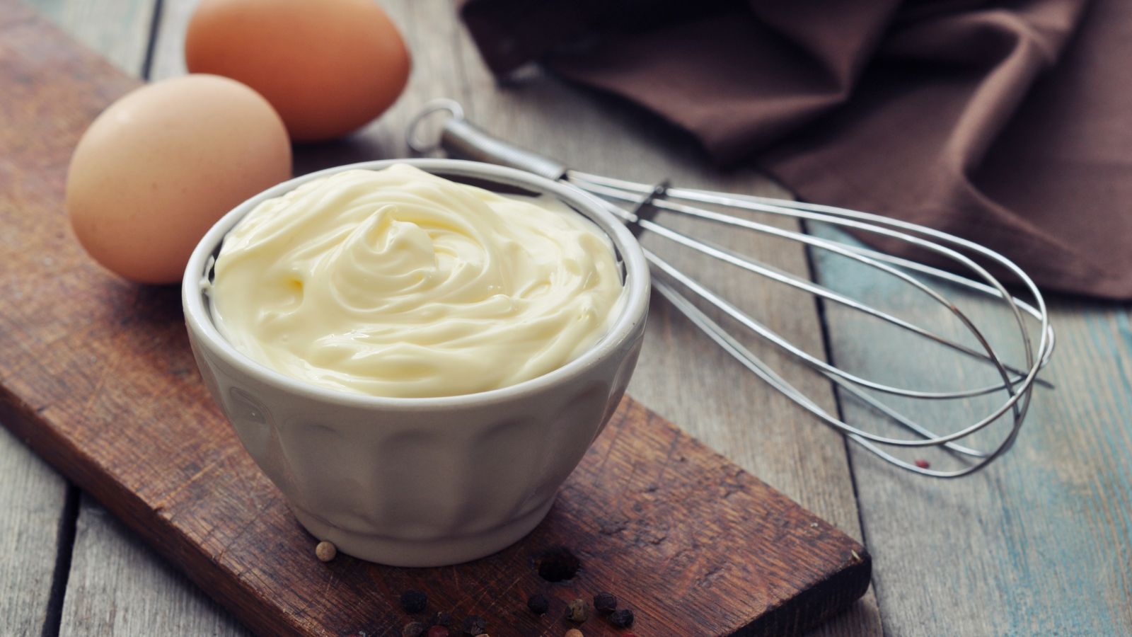 Fresh mayo in white bowl with eggs and whisk on wooden board.
