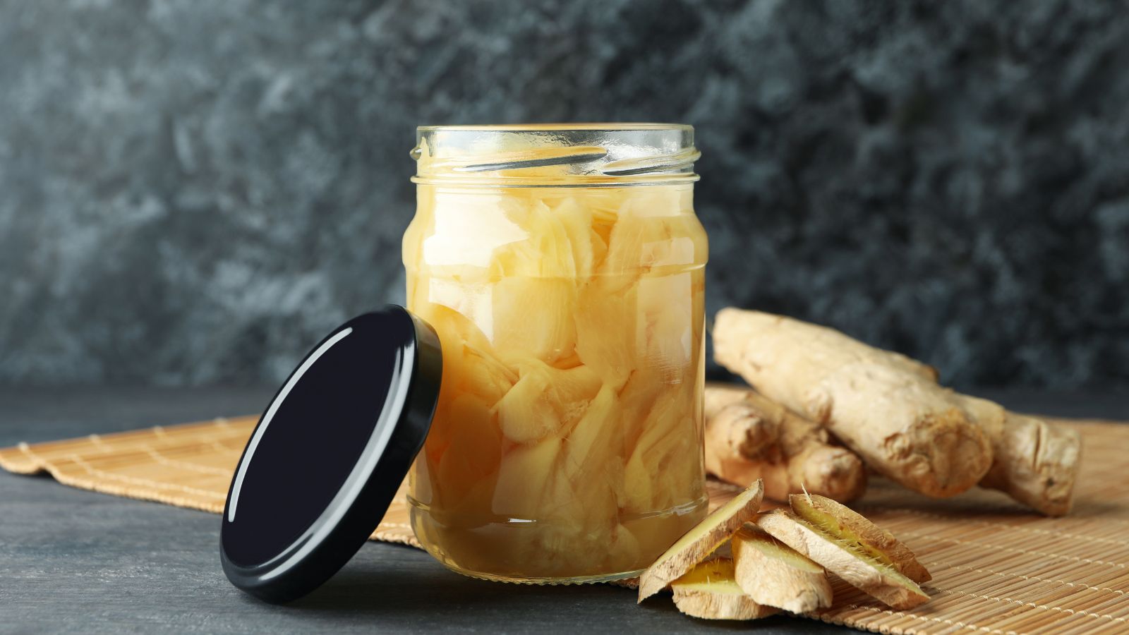 Pickled ginger slices in glass jar with fresh root on bamboo mat