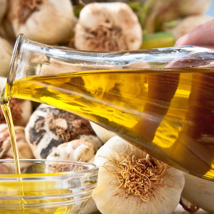 Hand pouring golden vegetable oil from a glass jug into a bowl over garlic.