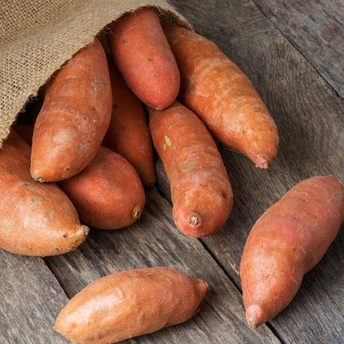 Burlap sack spilling sweet potatoes on wooden surface.