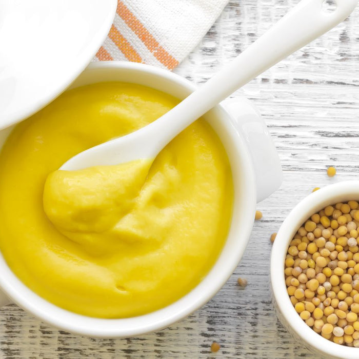 Yellow mustard in white bowl with mustard seeds beside it.