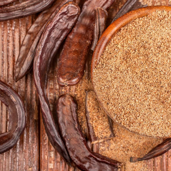 Ground carob powder in a wooden bowl with dried carob pods on a table.