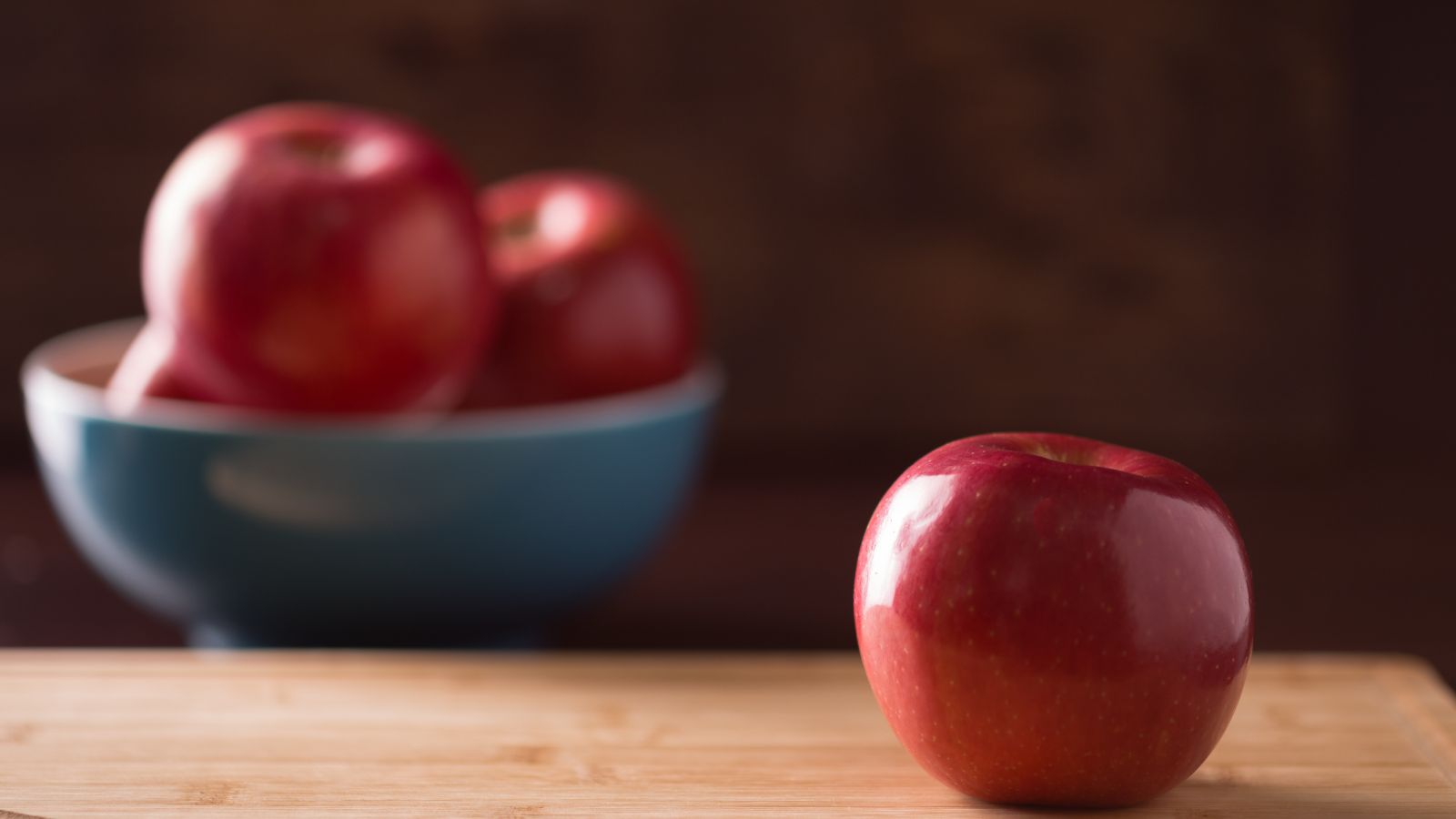 Shiny red apple on wooden surface with bowl of apples in background.