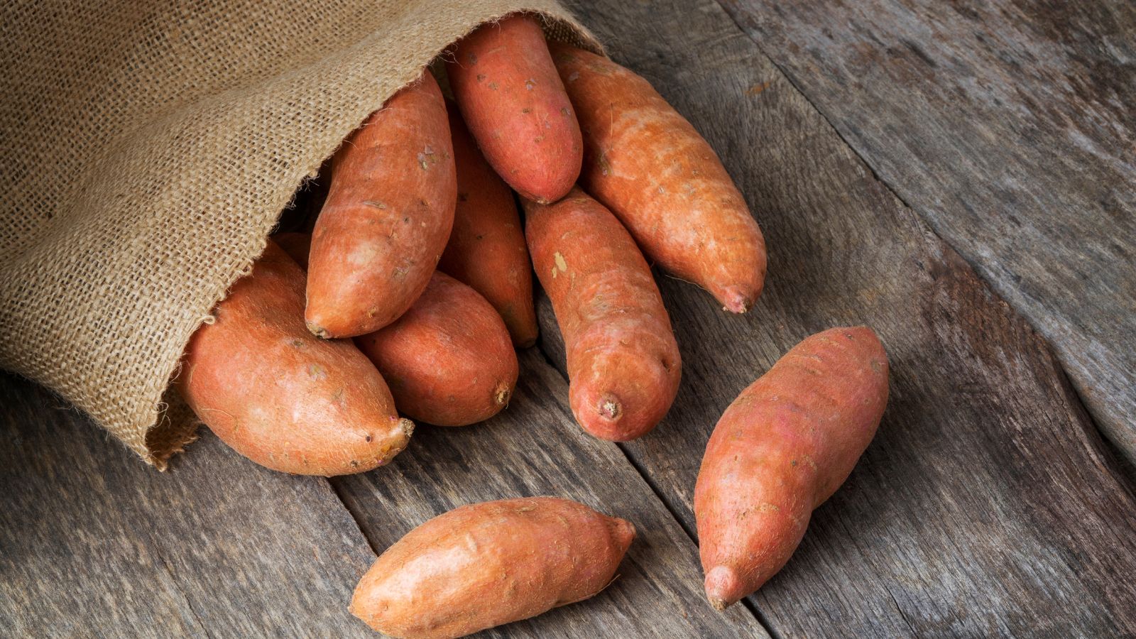 Burlap sack spilling sweet potatoes on wooden surface.