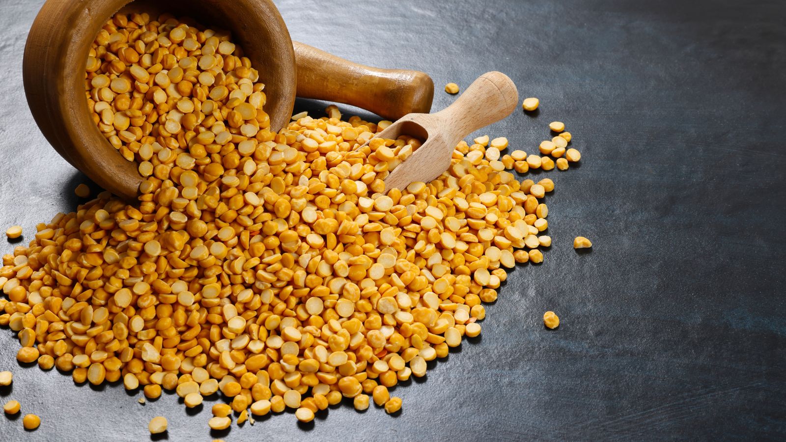 Yellow lentils spilled from a wooden bowl onto a dark surface.