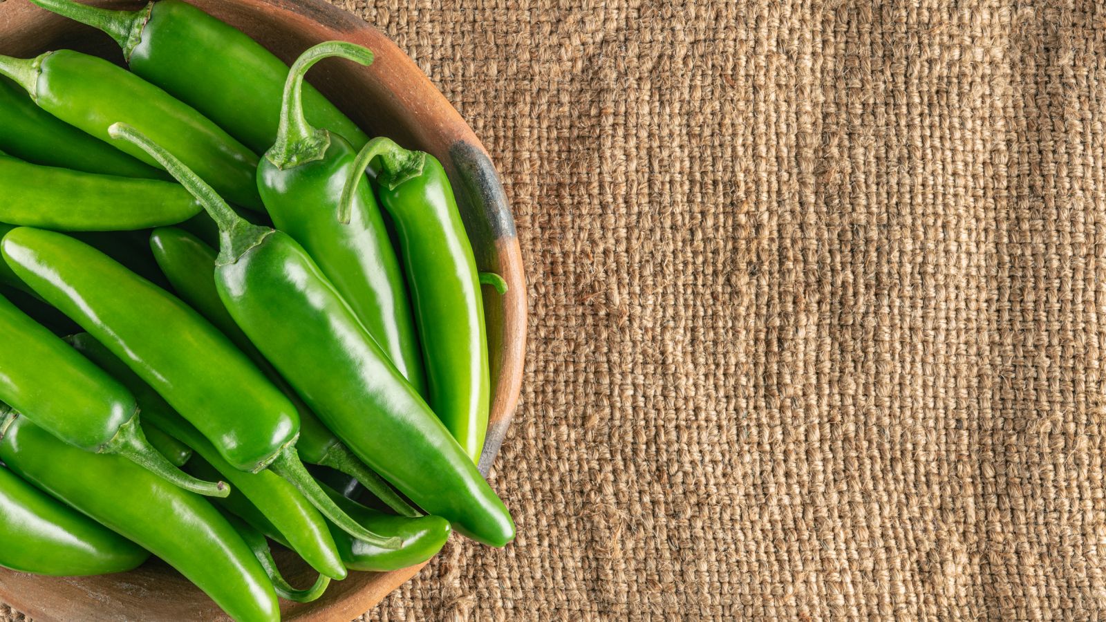 Fresh green jalapeno peppers in wooden bowl on burlap background