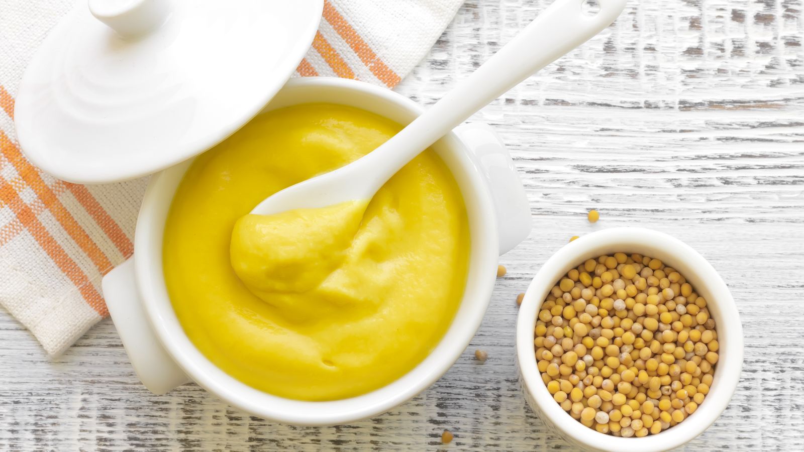 Yellow mustard in white bowl with mustard seeds beside it.