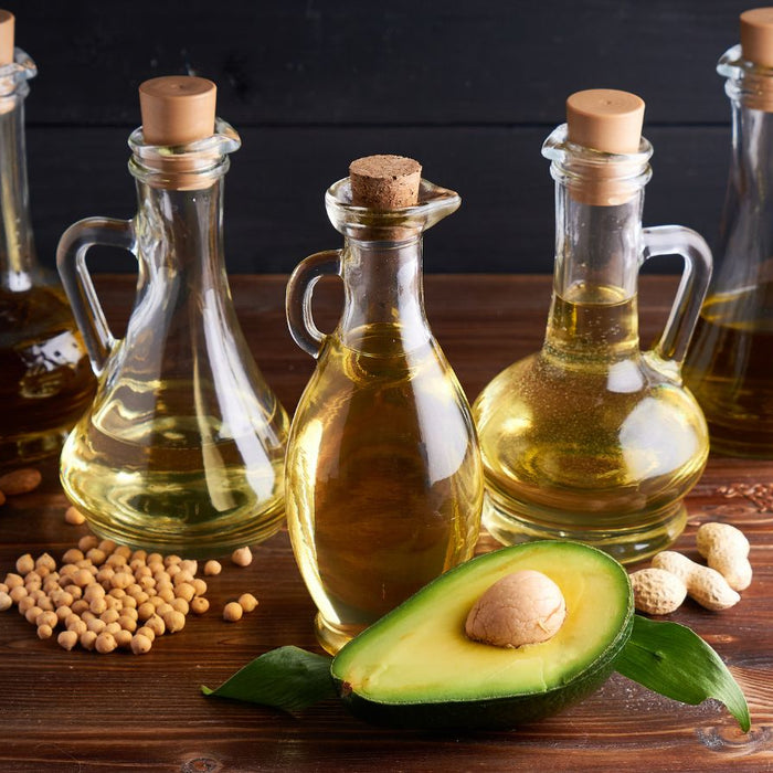 Assorted vegetable oils in glass bottles with avocado on wooden table.