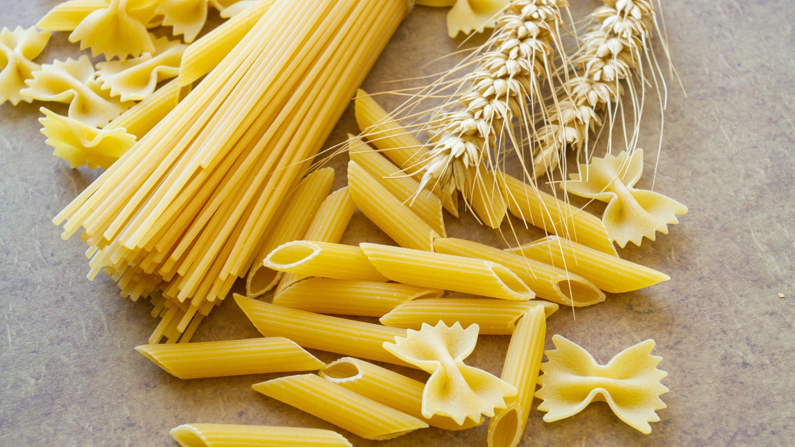 Various pasta shapes with wheat on a brown surface background.