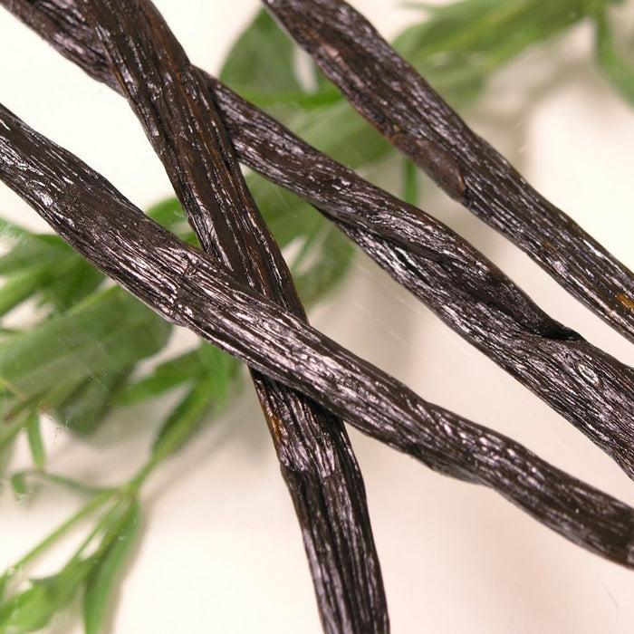 Three dark vanilla bean pods on white surface with green leaves.