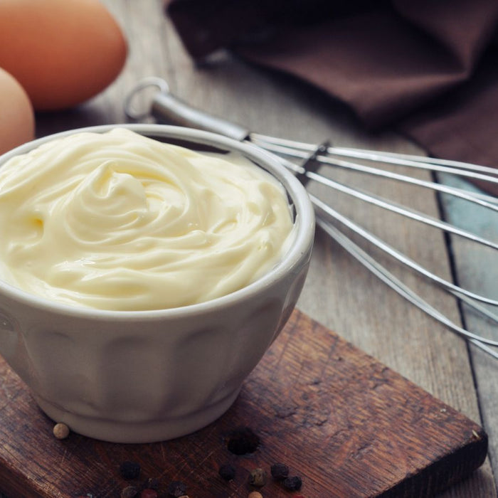 Fresh mayo in white bowl with eggs and whisk on wooden board.