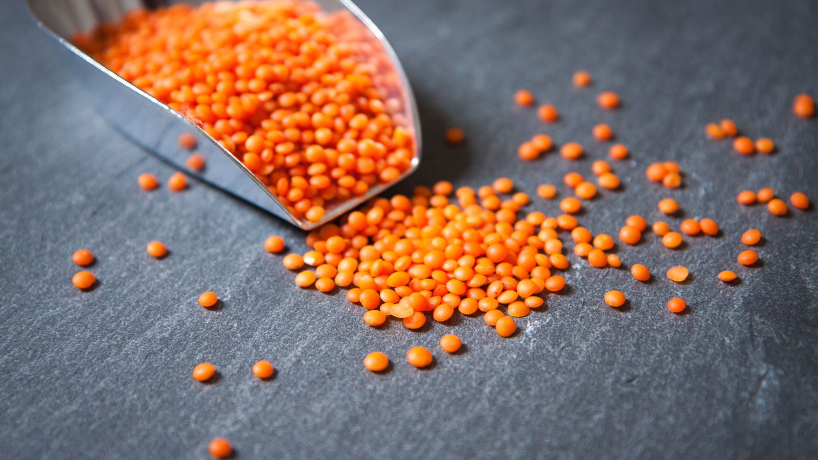 Red lentils spilling from silver scoop on dark stone surface