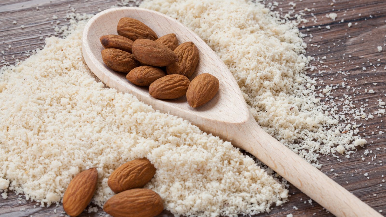 Wooden spoon with almonds rests on pile of almond flour on rustic wood.