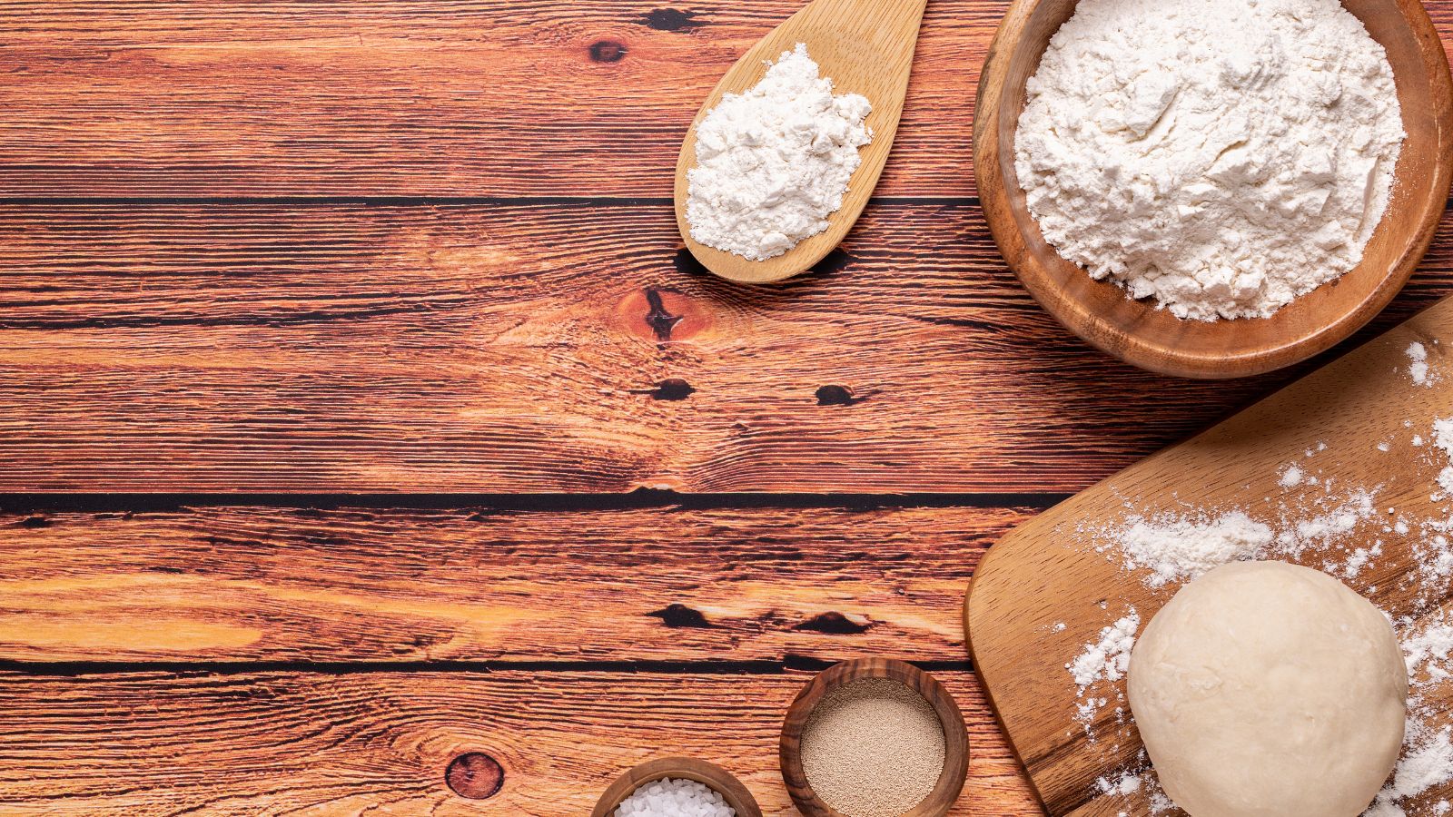Flour, yeast, and dough ball on wooden table with utensils.