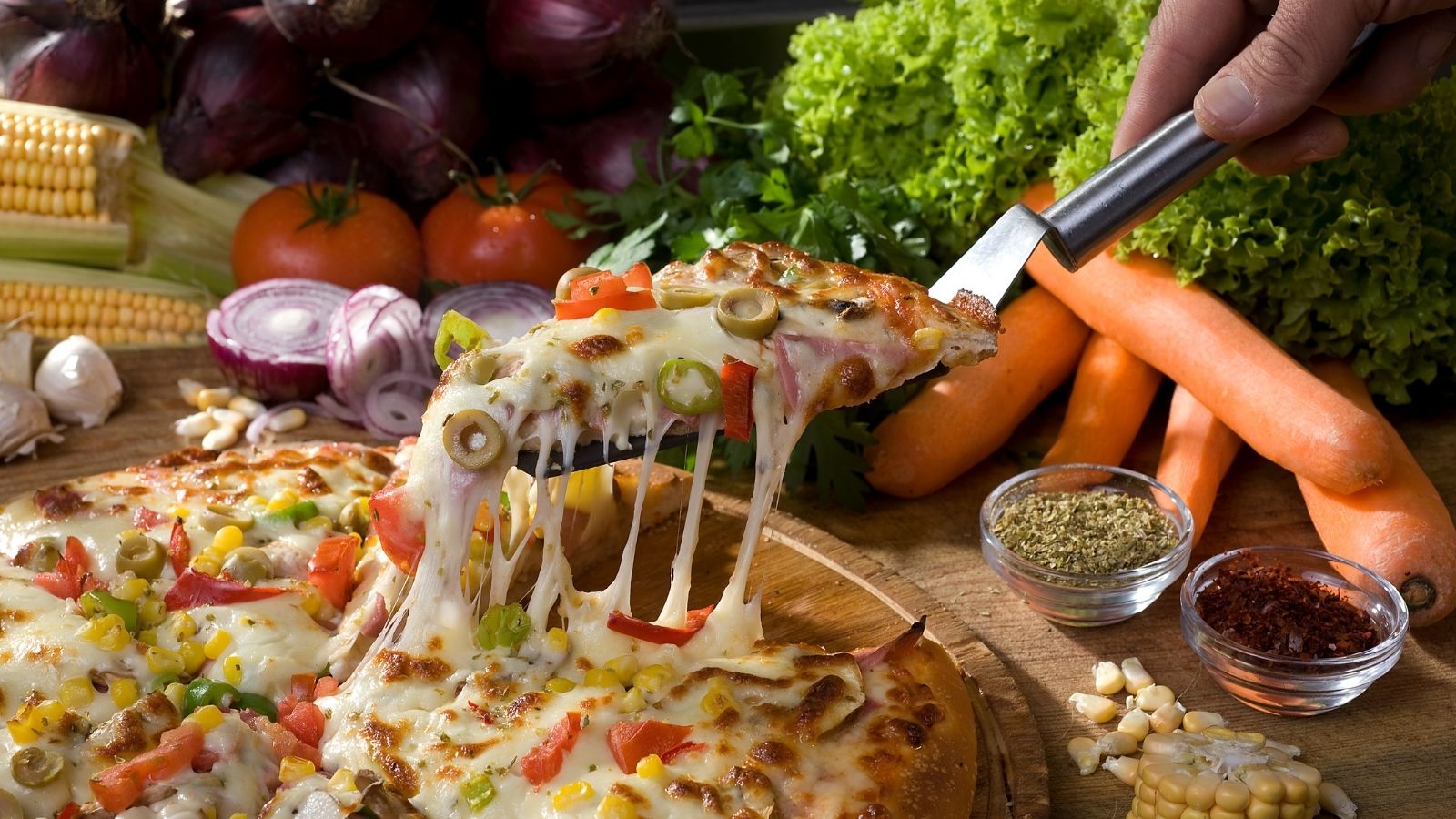 Slice of veggie pizza being lifted surrounded by fresh vegetables.