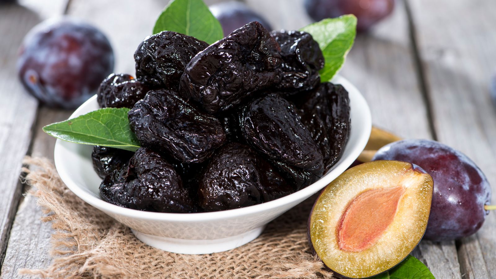 Ceramic bowl holds dark prunes beside fresh plum halves and green leaf.