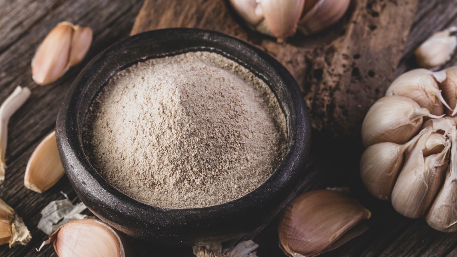 Bowl of garlic powder surrounded by garlic cloves on table.
