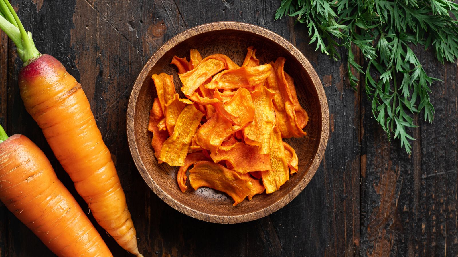 Wooden bowl filled with crispy carrot chips next to fresh carrots.