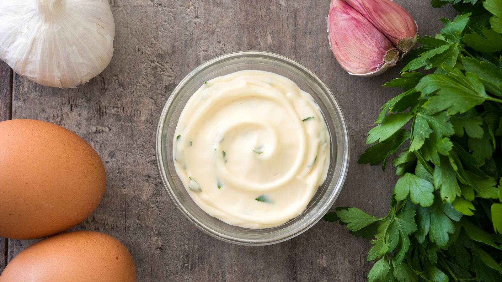 Garlic mayo sauce with herbs, eggs, garlic, and parsley on wood.