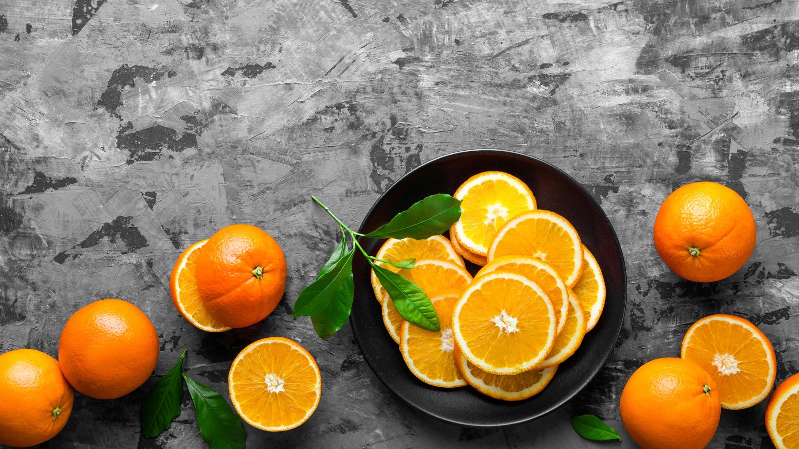Black plate of orange slices with whole oranges and green leaves.