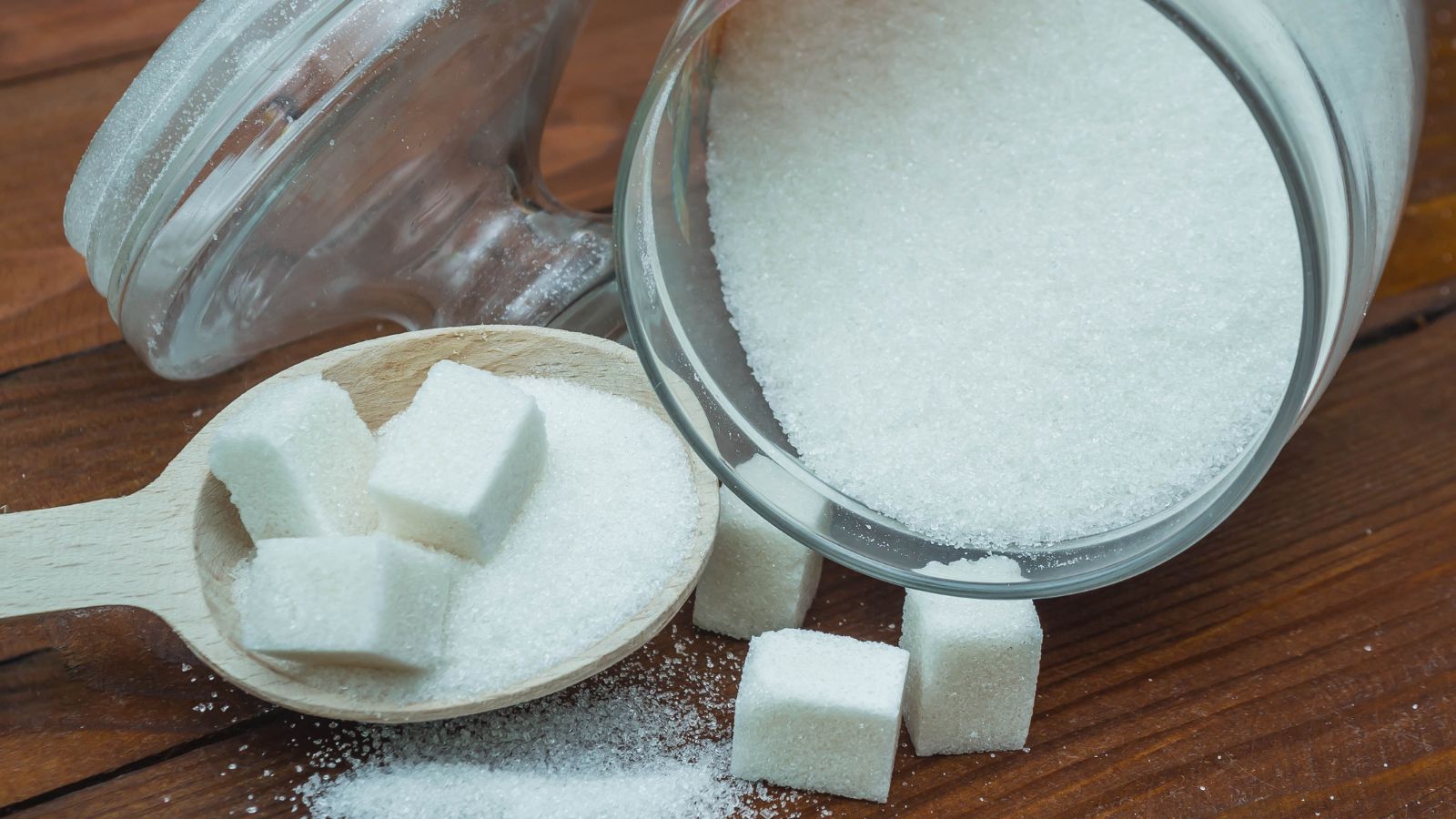Jar of white sugar with cubes on wooden spoon and tabletop.