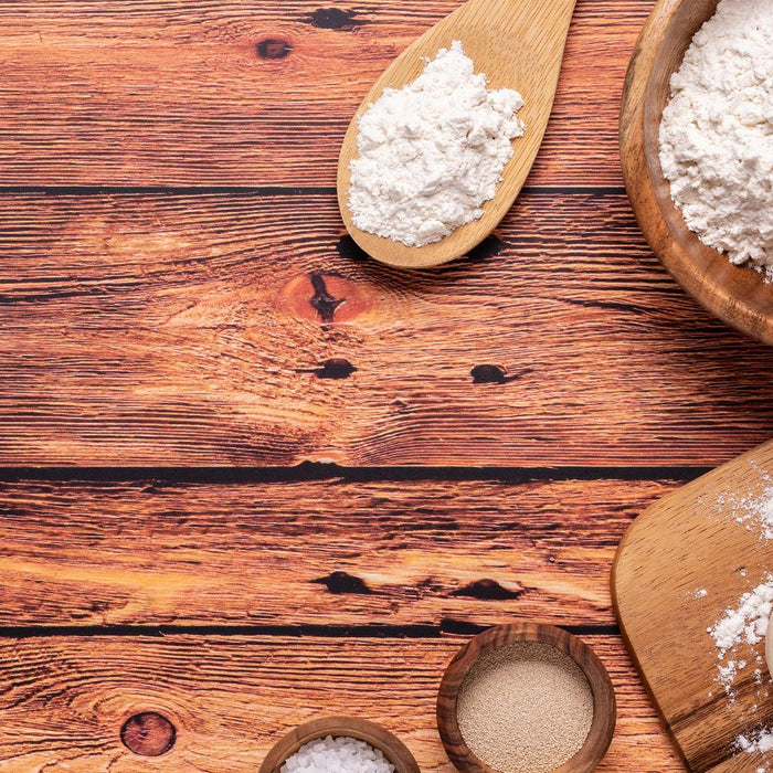 Flour, yeast, and dough ball on wooden table with utensils.