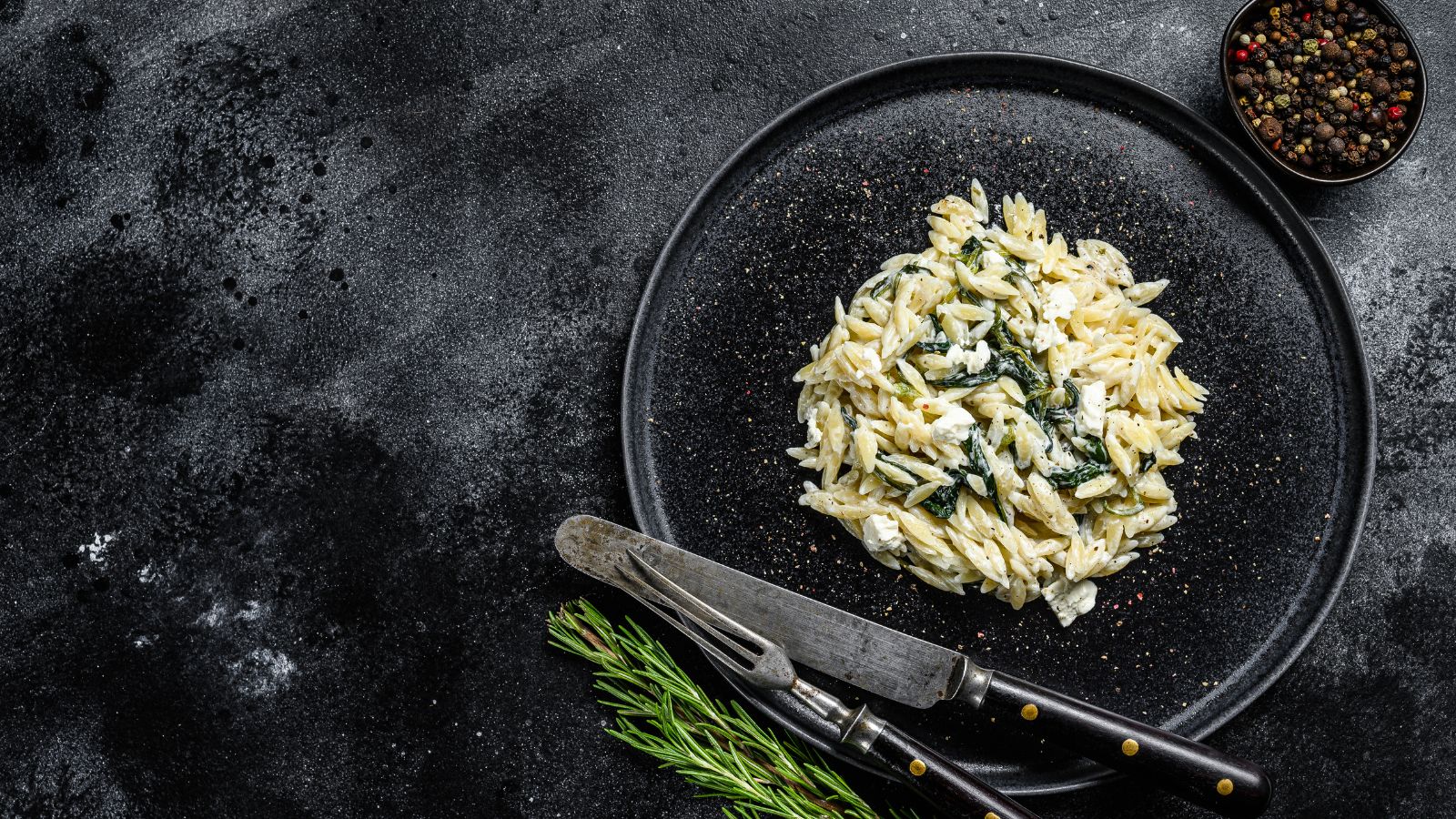Orzo pasta with spinach and feta on a black plate with herbs.