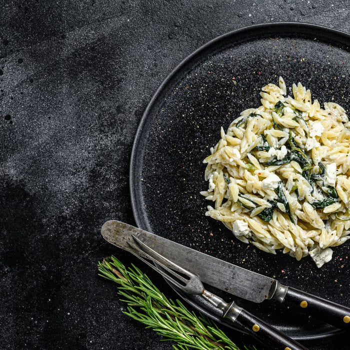 Orzo pasta with spinach and feta on a black plate with herbs.