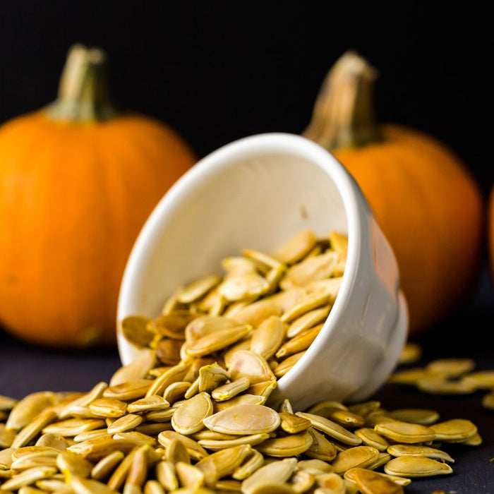 White bowl of pumpkin seeds spilling in front of whole pumpkins.