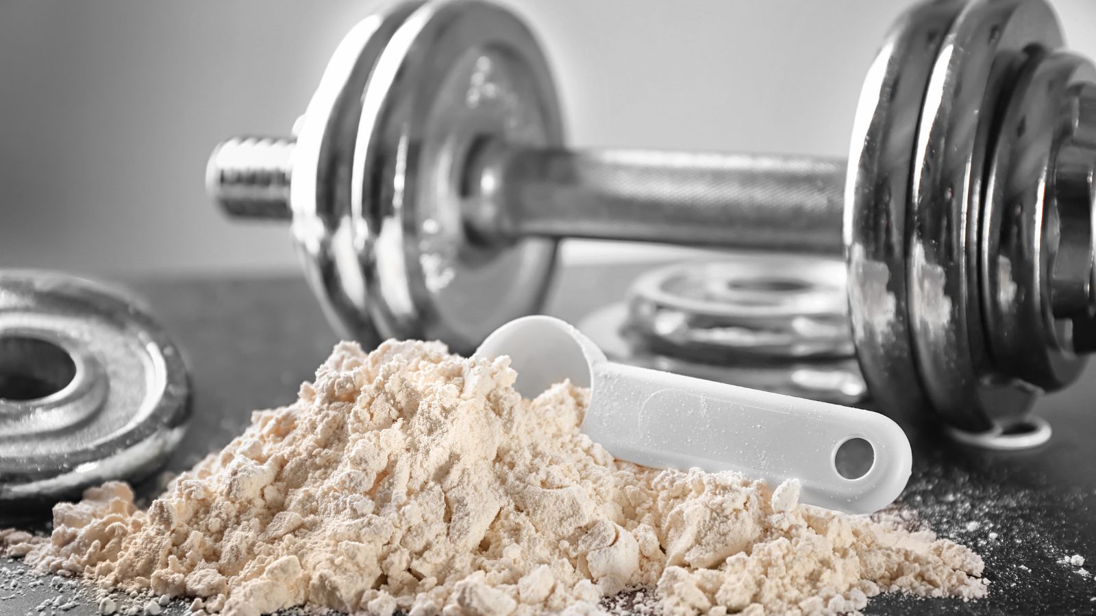 Protein powder with a white scoop placed near a metallic dumbbell