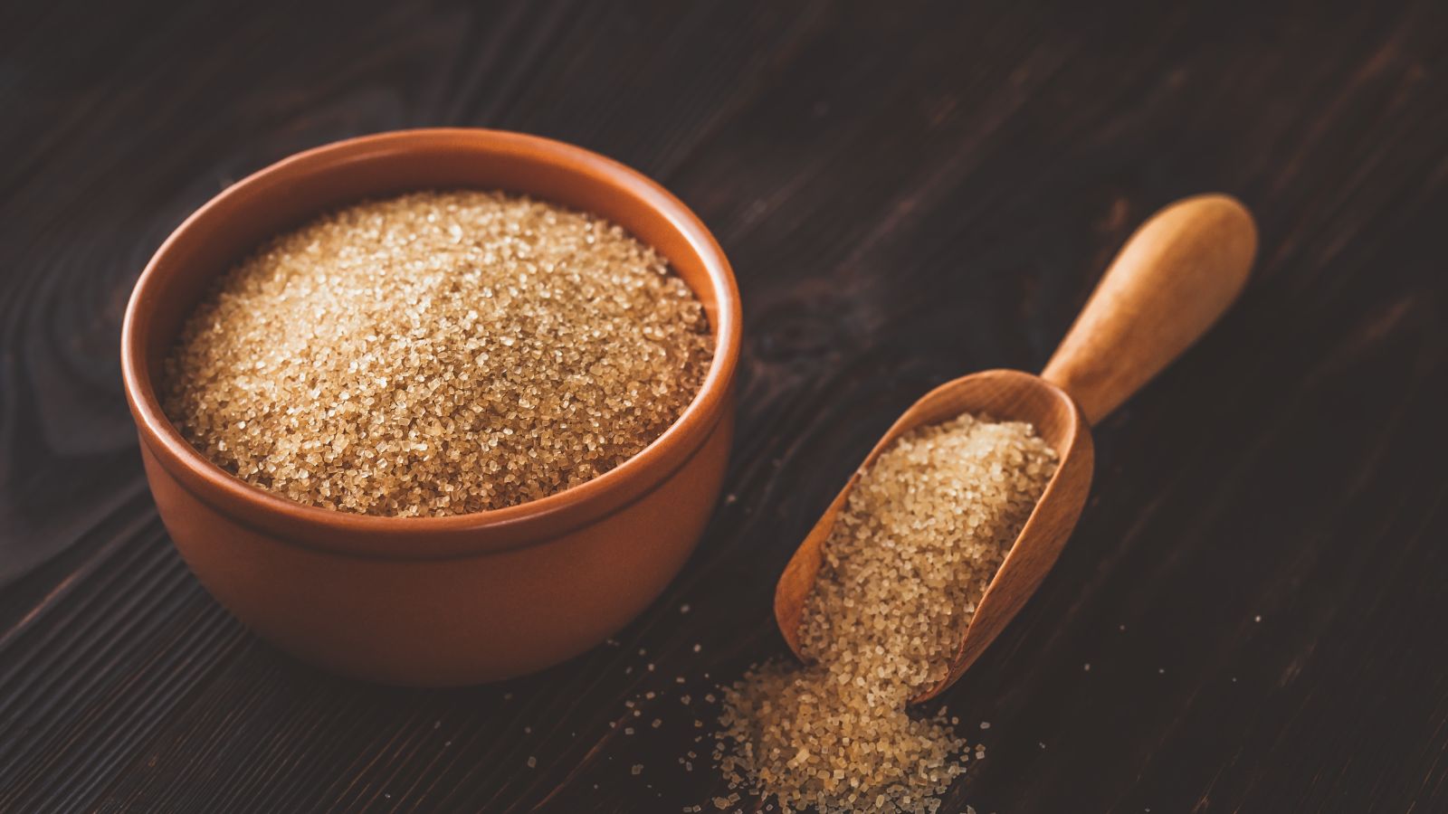 Raw brown sugar in bowl with wooden scoop on dark table.