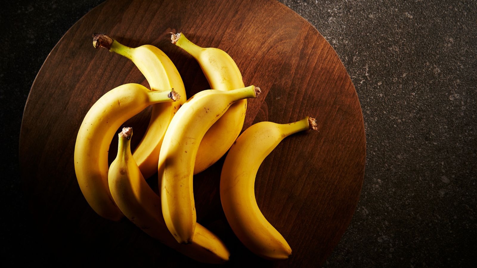 Six ripe bananas laid on a round wooden board over dark surface.